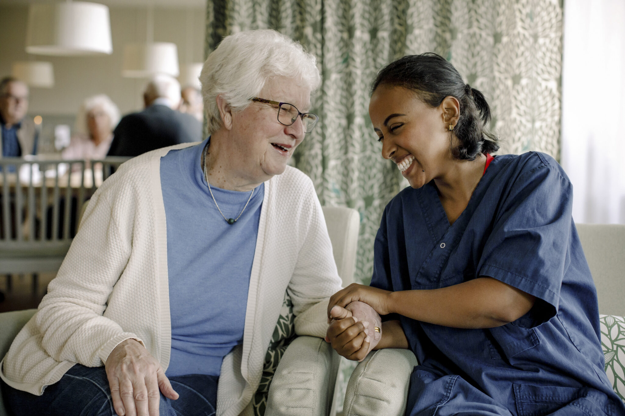 A health care worker sits side by side with an elderly woman and they share a laugh and hold hands